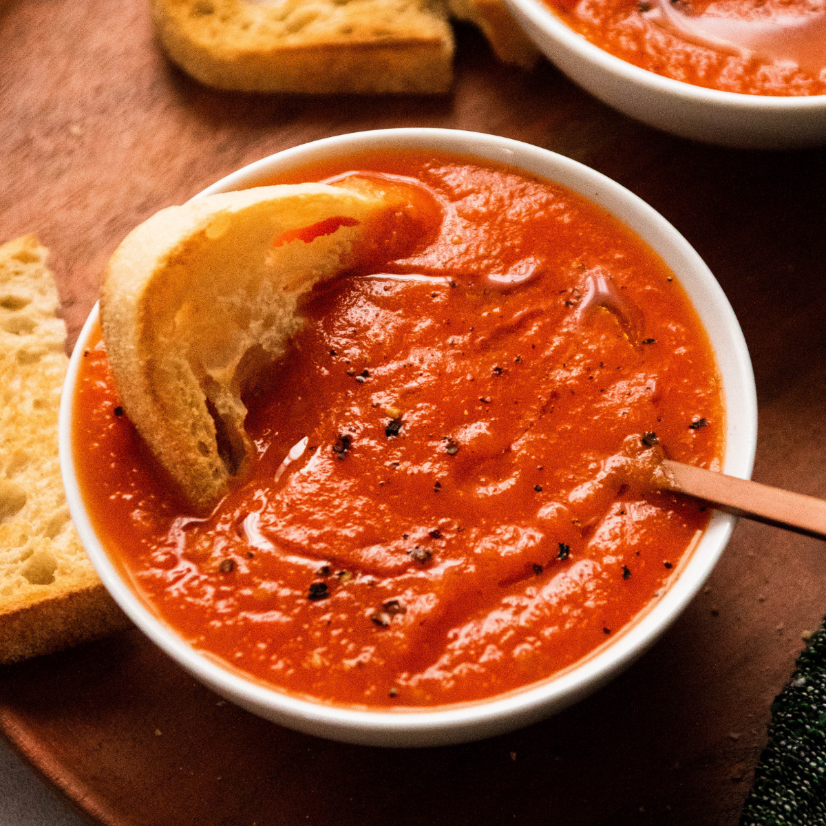 Tomato soup in a white bowl, with a copper spoon and a slice of bread.