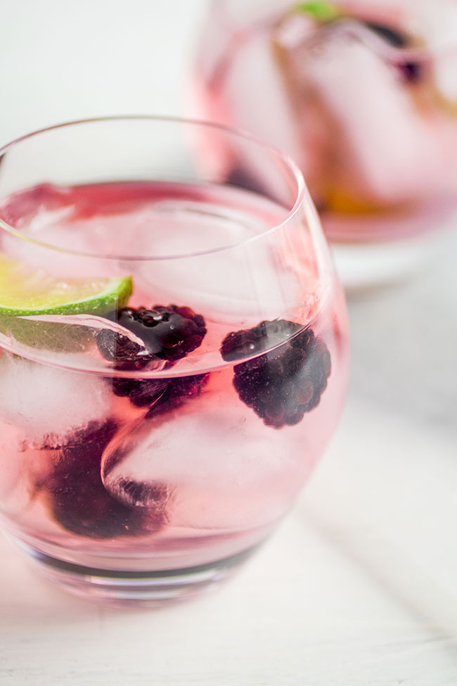 Two cocktail glasses filled with vodka tonics, fresh blackberries, and lime wedges on a white background.