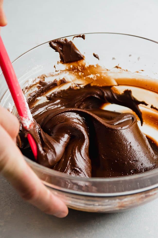 Man's hand stirring chocolate frosting with a red spatula.