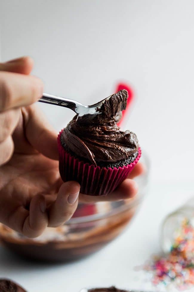 Man\'s hand using a spoon to frost a chocolate cupcake.