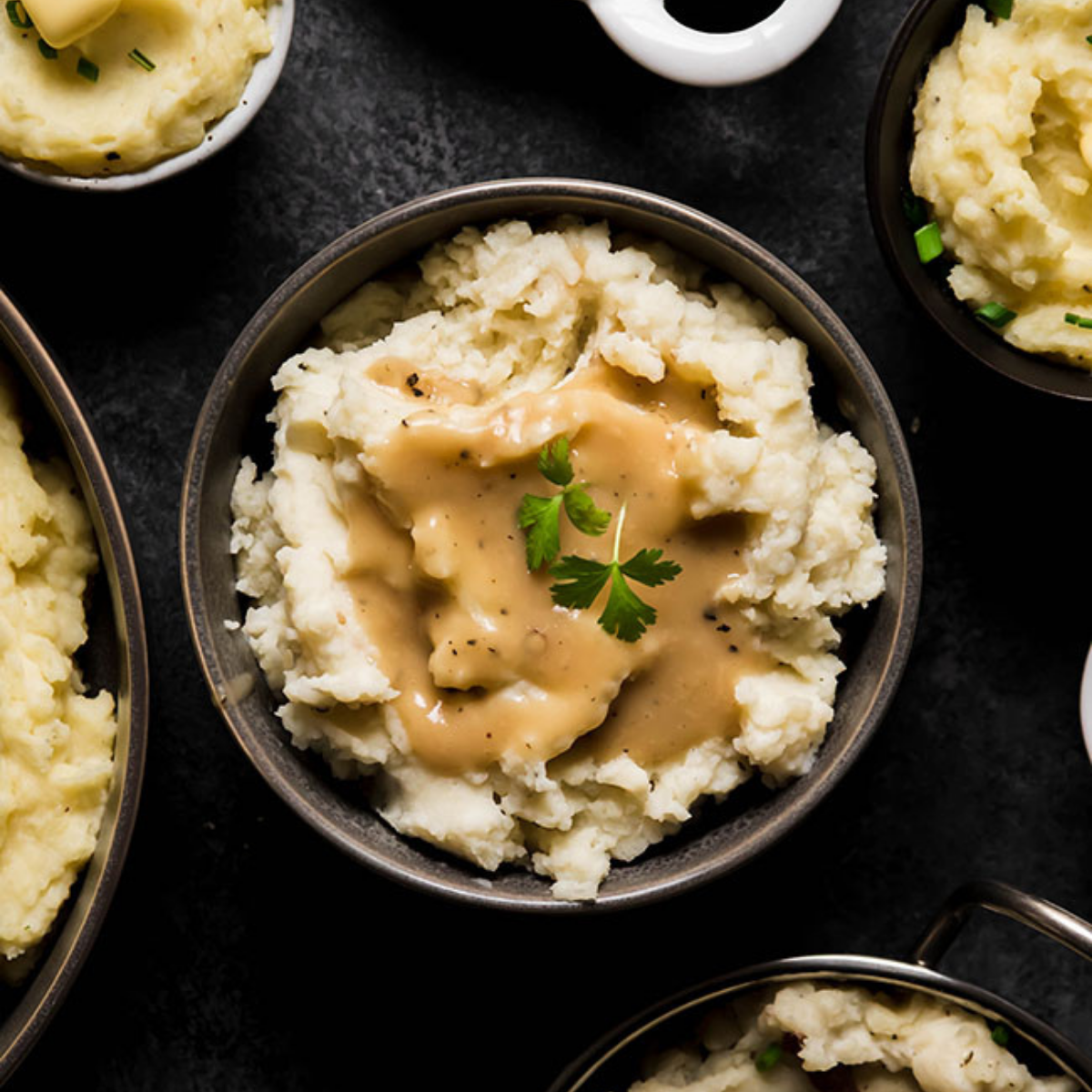 Several small bowls of mashed potatoes on a dark table with a variety of toppings like gravy, herbs, and melted butter.