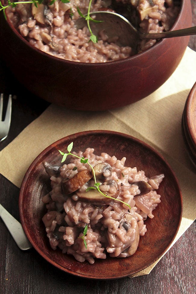 Light pink risotto on a dark wooden plate with a sprig of fresh thyme, next to a wooden serving bowl.