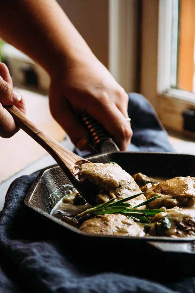 Hands using a wooden spoon to lift a piece of chicken out of a skillet.