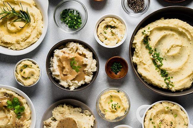 Aerial view of multiple small bowls full of mashed potatoes on a white table, each with various toppings.