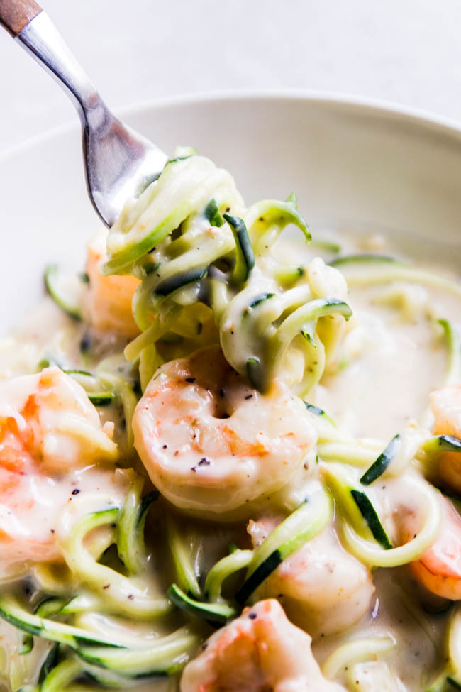 Fork lifting zucchini noodles with alfredo sauce and shrimp out of a white bowl.