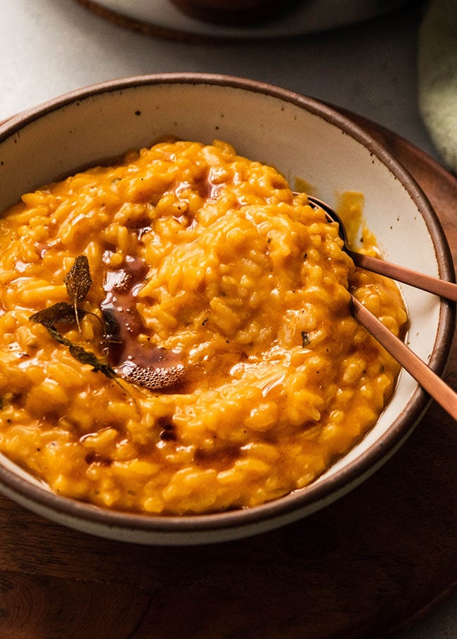 Two copper spoons tucked into a speckled white bowl filled with bright orange butternut squash risotto.